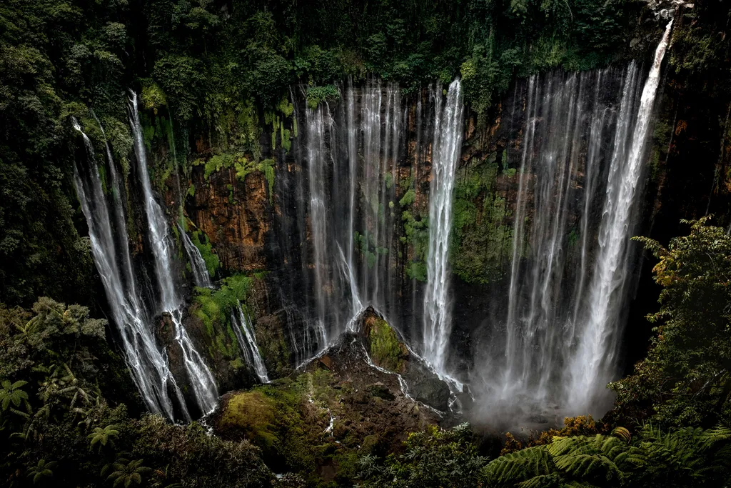Majestic Tumpak Sewu waterfall in Indonesia, with countless streams cascading down a lush green cliff face into a misty basin.